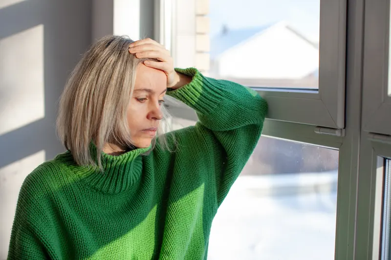 Frau steht am Fenster und fasst sich an die Stirn
